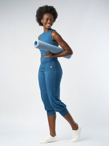Smiling woman in blue workout attire carrying a rolled-up light blue yoga mat on white background.