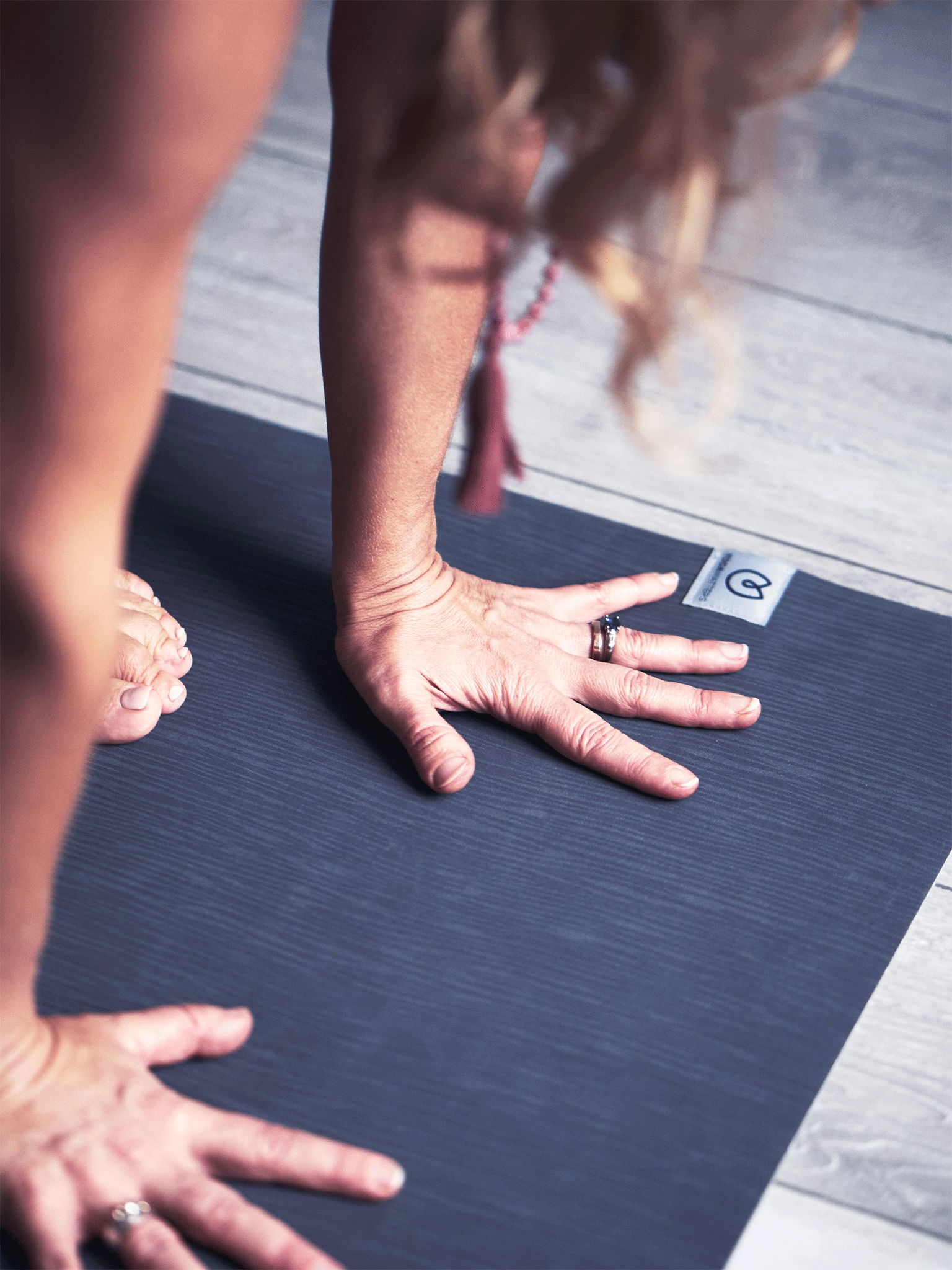 Woman practicing yoga with close-up on hands in downward-facing dog position on a dark grey yoga mat, featuring a visible logo, shot from the side on a wooden floor background.
