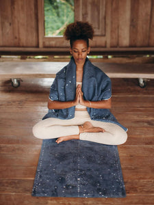 Woman practicing yoga in calm indoor setting on dark blue galaxy print yoga mat, wearing cream leggings and blue shawl, in a serene seated pose expressing mindfulness and tranquility.