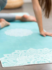 Turquoise blue yoga mat with mandala pattern, non-slip textured surface, eco-friendly material, image captured from side with partial view of female practitioner in a home setting.