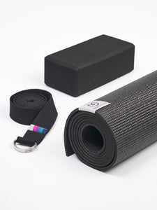 Black textured yoga mat rolled up with carrying strap and foam block on white background, side angle view.