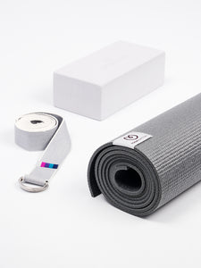 Gray textured yoga mat rolled up with branded tag visible, accompanied by white yoga block and gray carrying strap, shot from above on a white background.