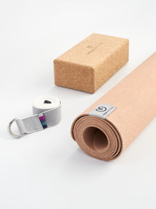 Cork yoga block, rolled terracotta yoga mat, and gray yoga strap with metal buckle on white background - side view.