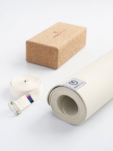 Beige yoga mat with brand logo, cork yoga block, and white strap with metal buckle shot from the front on a white background.