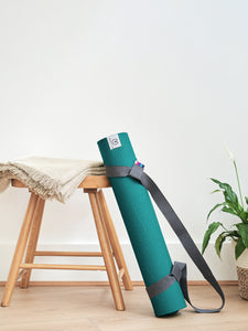 Green textured yoga mat with carrying strap leaning against wooden stool in a minimalistic room, side view displayed with a small potted plant in background.