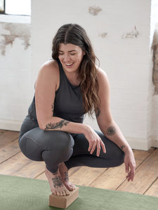 Woman practicing yoga on a green yoga mat with a cork block, wearing gray activewear in a bright studio space.