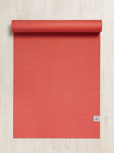 Red textured yoga mat partially rolled up lying flat on a wooden floor, front view, with visible brand logo on the bottom right corner.