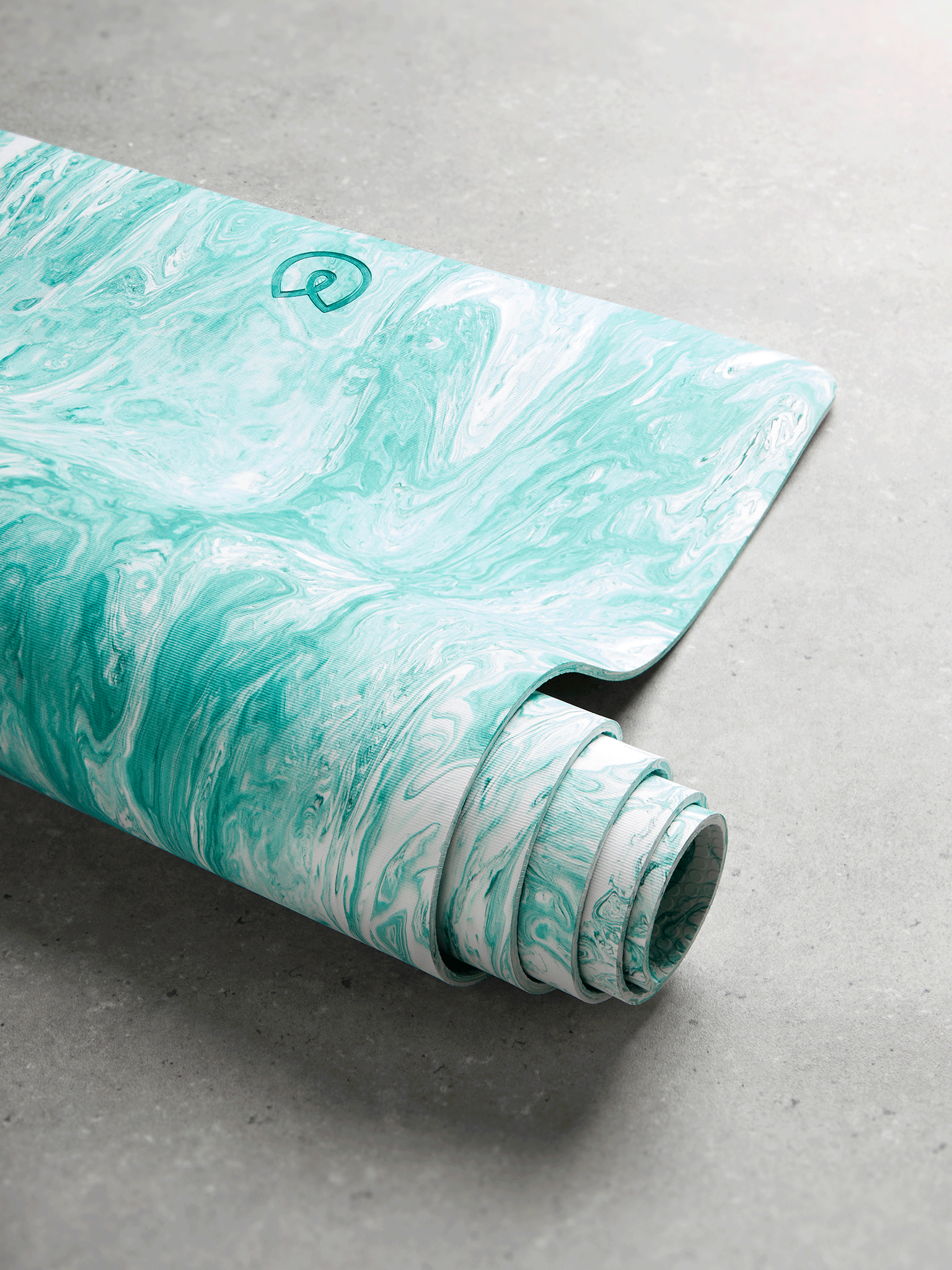 Luxury turquoise marbled yoga mat partially unrolled on grey concrete floor, side perspective, textured non-slip surface visible, exercise and wellness accessory