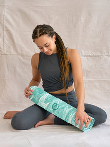 Woman in gray yoga attire rolling teal patterned yoga mat from side perspective against neutral backdrop
