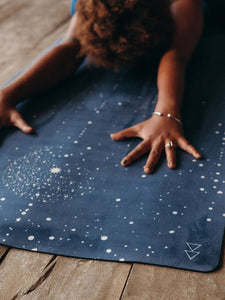 Navy blue yoga mat with white celestial pattern, top view, person practicing yoga with hands on mat, wooden floor background, non-slip textured surface.