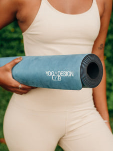 Side view of a person holding a blue YOGADESIGN LAB yoga mat, eco-friendly material, non-slip texture, perfect for exercise and fitness routines, outdoor greenery background.