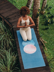 Woman practicing yoga in nature on a blue ombre yoga mat with circular pink design, shot from above showing peaceful outdoor setting