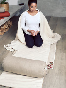 Woman meditating on beige yoga mat, side angle view, with matching bolster and meditation cushion visible, in serene home setting.