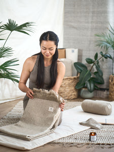 Woman practicing yoga at home unrolling taupe yoga mat with natural eco-friendly materials, indoor peaceful zen decor with plants, yoga accessories visible including bolster and essential oil.