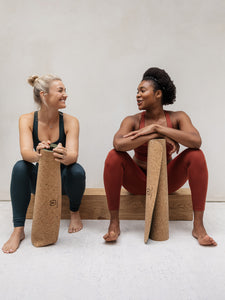 Two women in yoga attire sitting next to cork yoga mat with brand symbol, one in green top and black leggings, other in red bodysuit, engaging in conversation, neutral-toned background, indoor natural light setting.