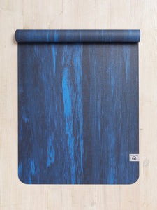 Blue textured yoga mat rolled partially with a visible logo shot from the front on a wooden floor background.