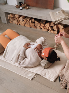 Neutral-toned yoga mat with visible brand label, woman lying on back with bolster pillow, dressed in orange and white, natural light, zen home atmosphere, relaxing with wine glass in hand.