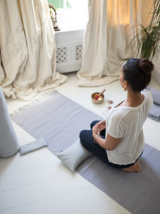 Woman in yoga practice pose on a gray yoga mat in a bright home interior, meditating in a serene setting with natural light, side view.