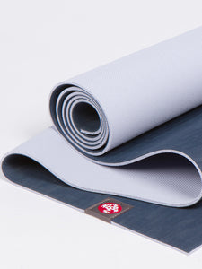 Gray yoga mat partially unrolled with visible Manduka brand logo, textured surface, shot from the side on a white background.