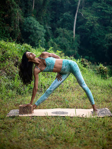 Woman practicing yoga in nature on beige yoga mat, side view, outdoor exercise, serene setting with trees, wearing blue patterned yoga pants and top, fitness lifestyle, meditation in forest environment.