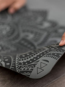 Close-up side view of a grey patterned yoga mat possibly from a recognizable brand, with a person's hands adjusting it on a wooden floor – ideal for mindfulness and fitness enthusiasts