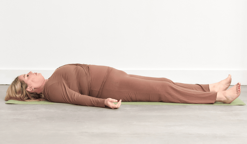 Person practising Corpse Pose (Savasana), lying on the back with the legs extended, arms resting alongside the body and palms facing upwards.