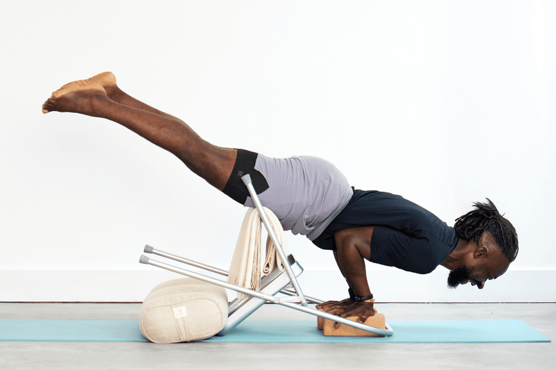 Person practising Peacock Pose (Mayurasana), balancing on the hands with the body and legs extended, using a yoga chair, bolster, folded blanket and yoga bricks for support.