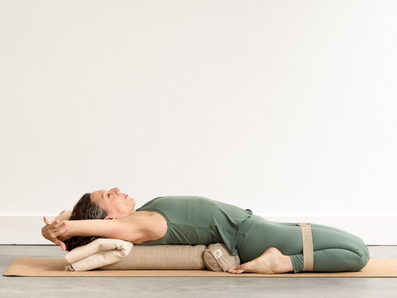 Person practising Reclined Hero Pose (Supta Virasana) using props, reclining backwards between the feet with the knees bent and the front of the body gently opening.