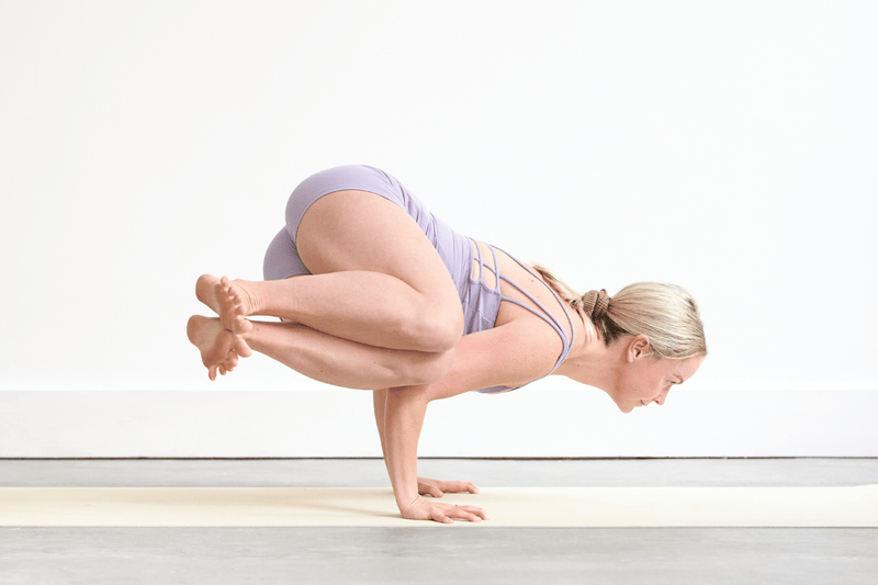 Person practising Side Crow Pose (Parsva Bakasana), balancing on both hands with the knees bent and legs lifted to one side.