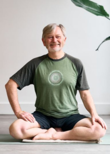 Man sitting on a yoga mat in a green and gray shirt with a logo, smiling.