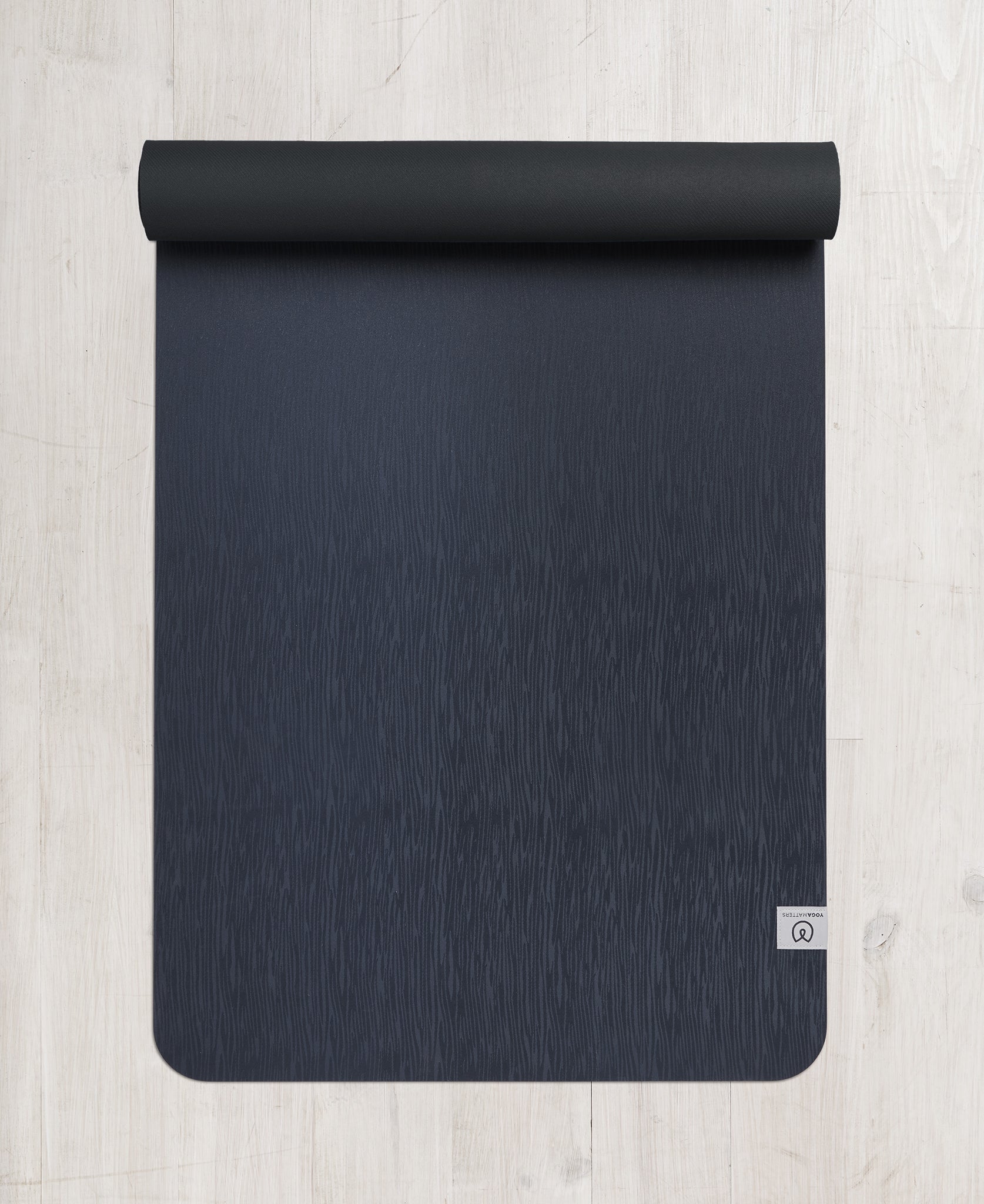 Dark navy blue premium yoga mat with textured surface, partially rolled up at the top, top view on a wooden floor background