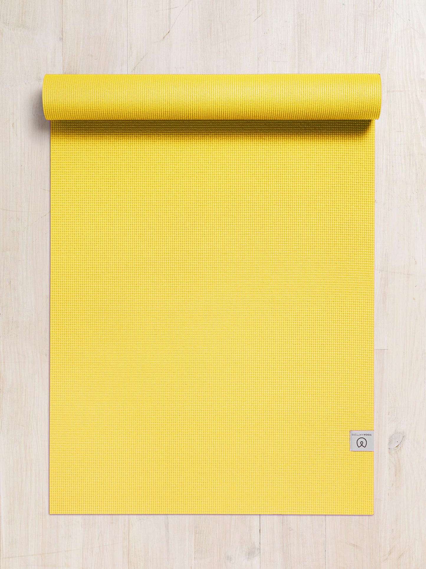 Bright yellow textured yoga mat partially rolled up at the top on a wooden floor, top view, with visible brand tag