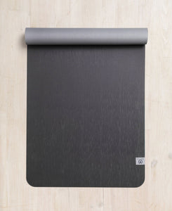 Top-down view of a rolled-up dark gray textured yoga mat on a wooden floor with visible brand logo on the bottom right corner.
