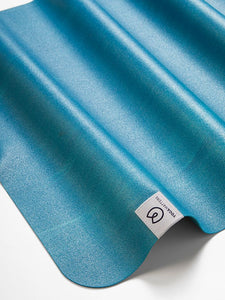 Blue gradient yoga mat with textured surface, featuring a prominent logo, shot from an angle showing the wavy profile of the mat.
