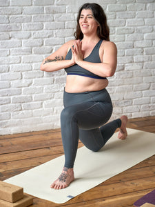 Woman in grey athletic wear practicing yoga on a cream-colored yoga mat, indoor natural light, wooden floor, white brick background, side perspective, yoga accessories(blocks) visible