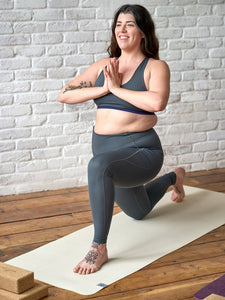 Woman practicing yoga in a tree pose on a beige yoga mat, front view, eco-friendly mat on wooden floor against a white brick wall, fitness and wellness concept, unidentifiable brand