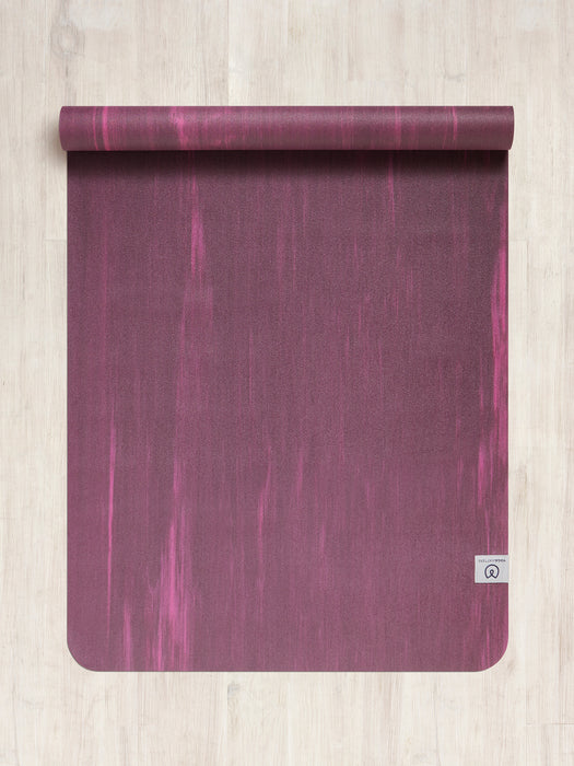 Top view of a textured deep purple yoga mat rolled out on a wooden floor, with a visible logo in the bottom right corner indicating a branded mat.