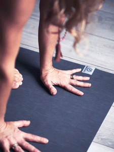Close-up angled view of a dark gray textured yoga mat with visible brand logo in a yoga studio, woman practicing a yoga pose with focus on hands and alignment on the mat.