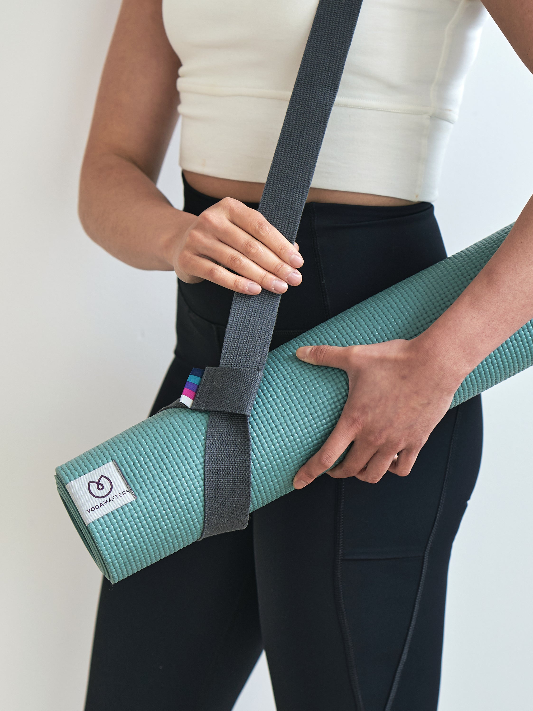 Close-up of a teal YogaMatters yoga mat rolled up and carried with a gray strap by a person in black leggings and a white top, shot from the side.