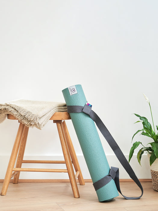 Teal yoga mat with carrying strap leaning against wooden stool in a home setting with neutral wall and green plant.