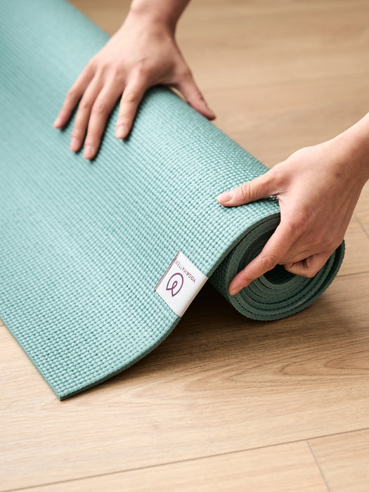 Textured teal yoga mat being rolled up on a wooden floor with hands, featuring a visible brand tag, eco-friendly non-slip yoga accessory in a close-up side shot.