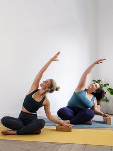 Two women practicing yoga side stretch pose on yellow mat with foam blocks, indoor yoga class, fitness and wellness lifestyle, side view.