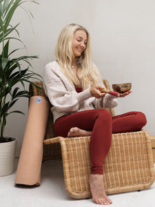 Blonde woman meditating with singing bowl, sitting beside cork yoga mat on rattan bench, indoor peaceful yoga setup, plant in background, natural yoga accessories, side view of yoga mat