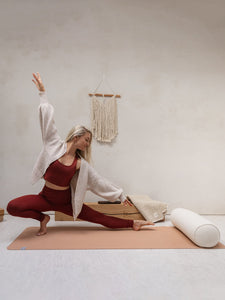 Woman practicing yoga in a side lunge pose on a peach-colored yoga mat, shot from the side with a rolled white yoga bolster, wooden props, and a wall-mounted macrame decoration in a minimalist setting.