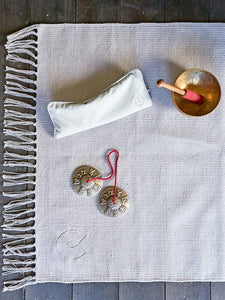 Top view of a grey textured yoga mat with fringe detail on hardwood floor, alongside a white towel, Tibetan singing bowl with striker, and Tibetan Tingsha cymbals.